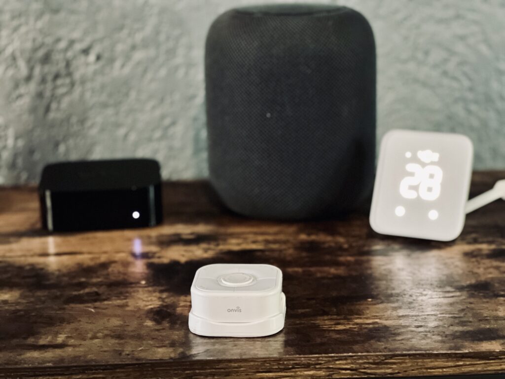 HS2 on wooden surface backed by a number of other Apple Home devices including Sensibo Air, the original HomePod, and SwitchBot Hub 2