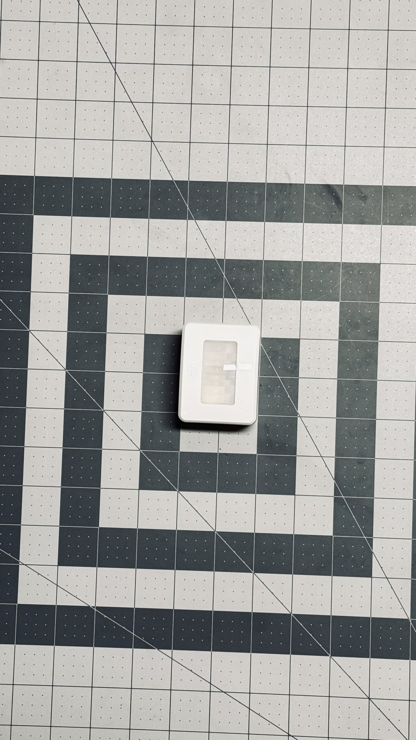 a small white rectangular cube with a smaller opaque rectangle on the face against a gray arts & crafts mat, the Onvis SMS2 Smart Motion Sensor