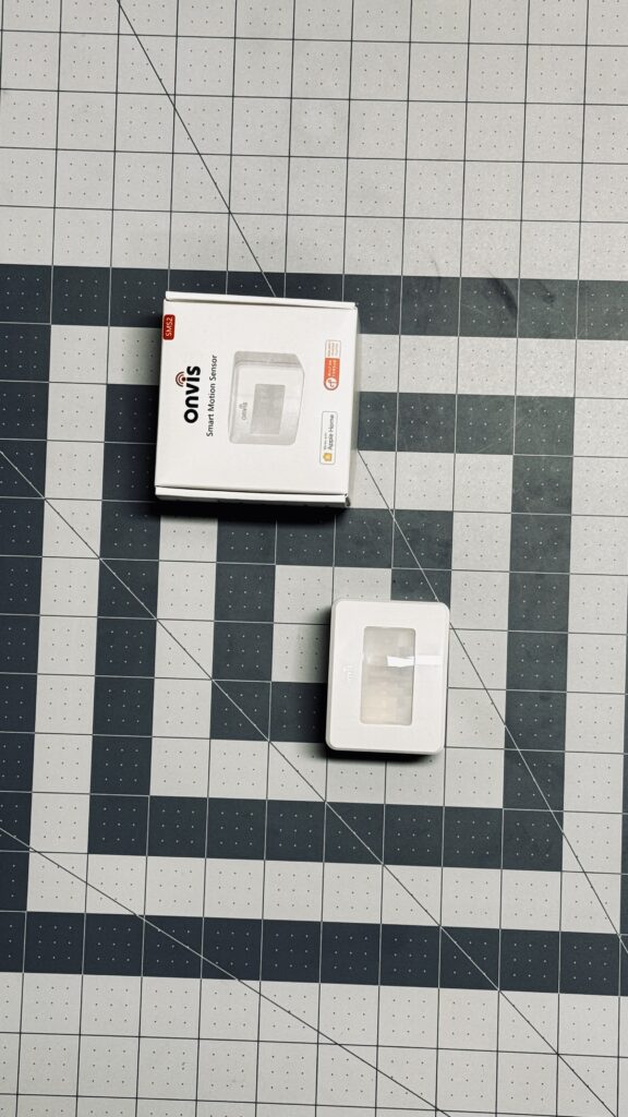 The Onvis SMS2 Smart Motion Sensor on a gray arts & crafts mat alongside its packaging